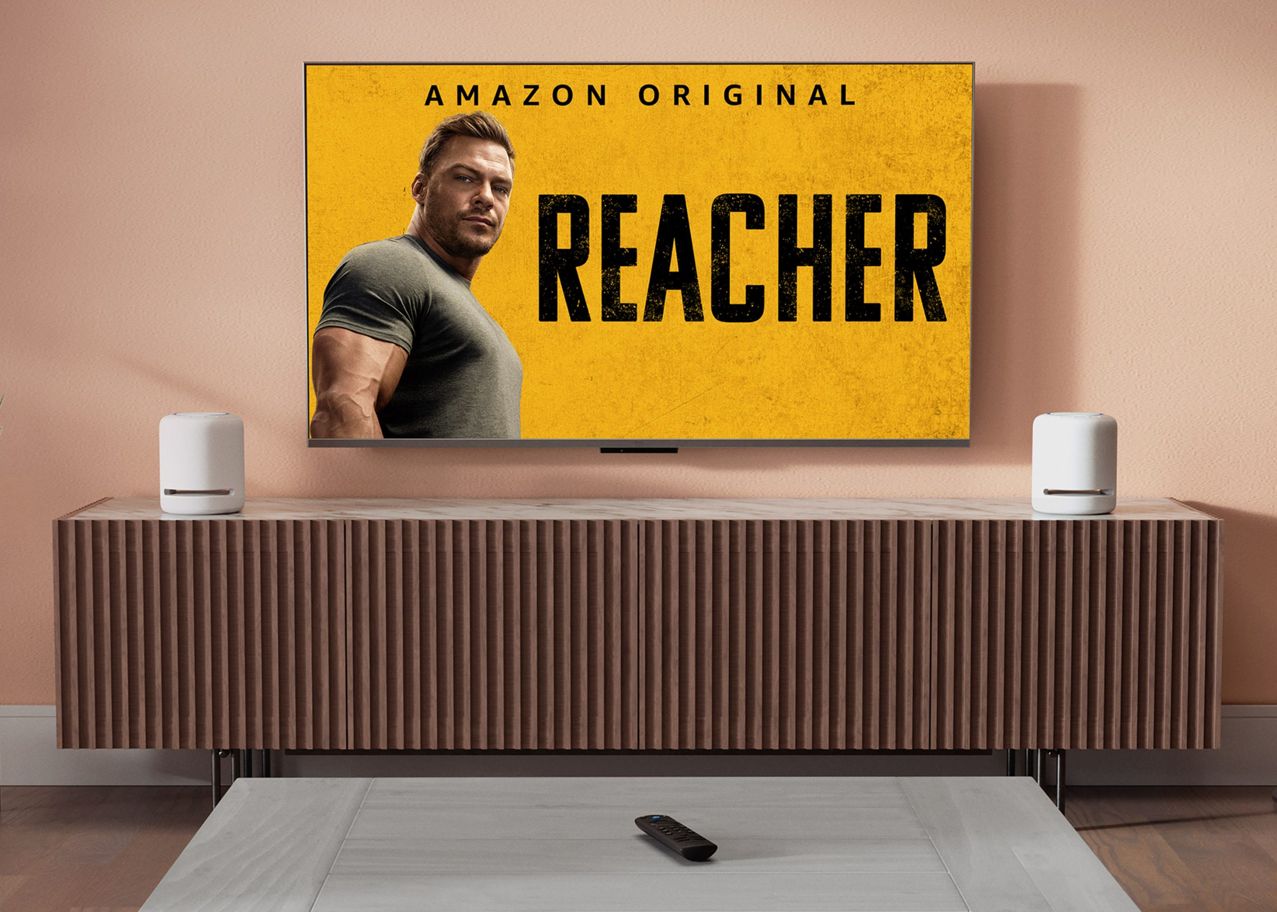 HDTV mounted on a wall in a modern living room showing Prime Video's "The Summer I turned Pretty" next to 2 Echo Studio: 7-inch wide and 8-inch tall cylindrical mesh speakers with flat bottoms and tops, and 4 raised buttons on the top.