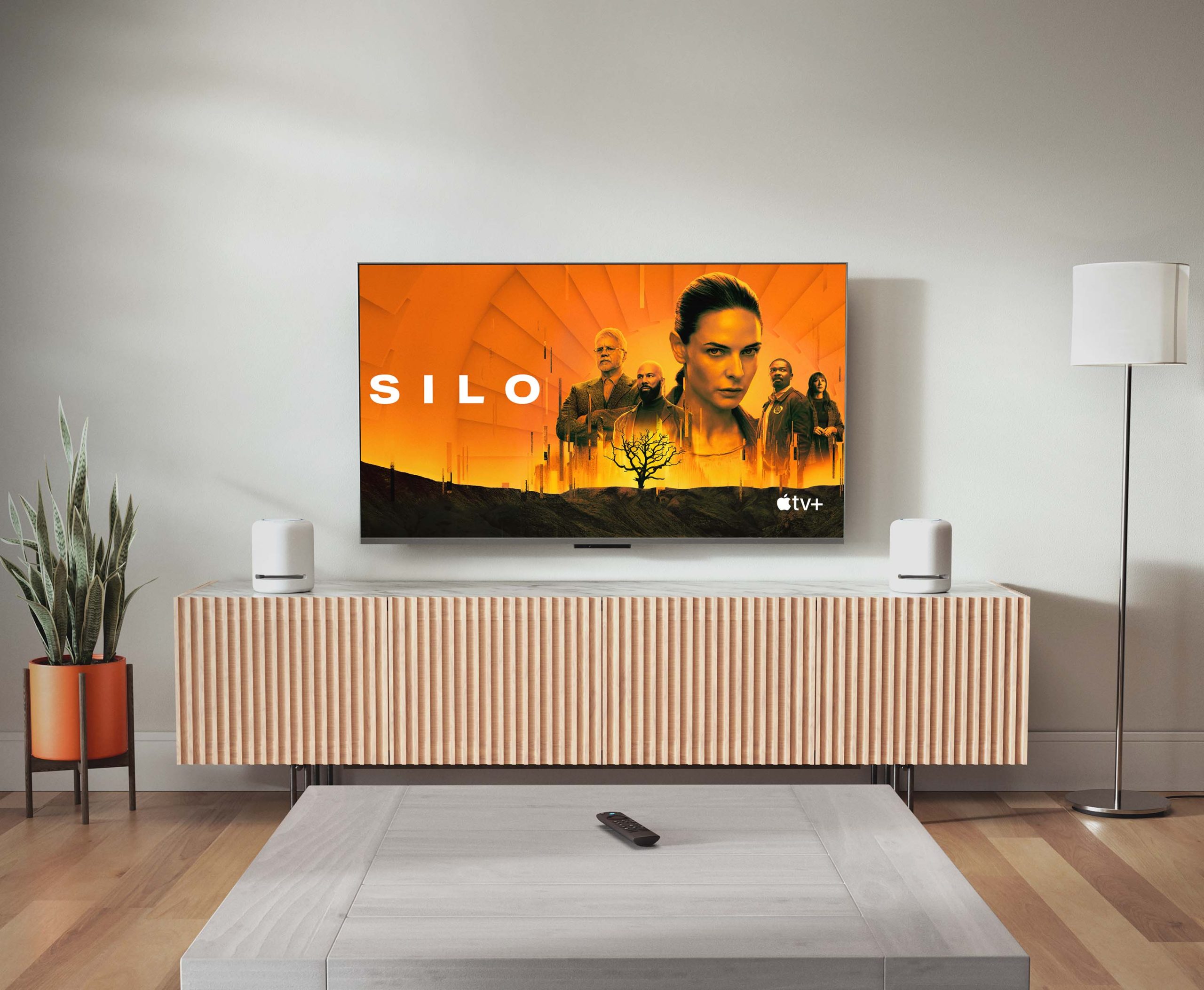 HDTV mounted on a wall in a modern living room showing Apple TV+ "Silo" next to 2 Echo Studio: 7-inch wide and 8-inch tall cylindrical mesh speakers with flat bottoms and tops, and 4 raised buttons on the top, sitting on a long table beneath it.